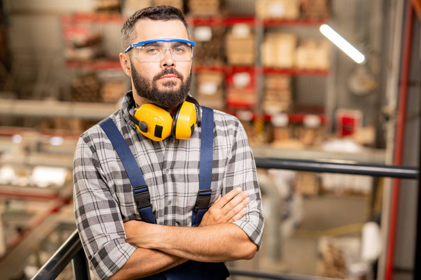 Trabajador de almacén serio en gafas protectoras y auriculares de pie delante de la cámara en el área de fábrica
 - Foto, Imagen