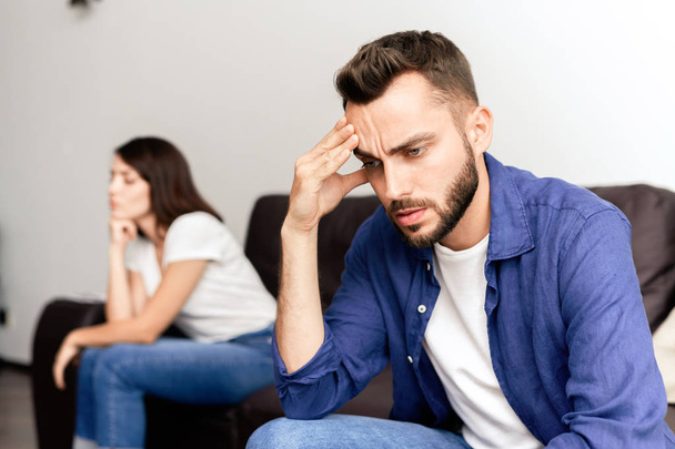 Upset young bearded man in blue shirt sitting on side opposite to girlfriend and rubbing forehead while suffering about fight - Photo, Image