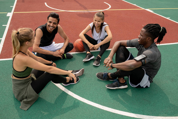 Onnelliset nuoret kulttuurienväliset ystävät keskustelevat viimeisestä koripallopelistä istuessaan kentällä harjoittelun jälkeen.
 - Valokuva, kuva