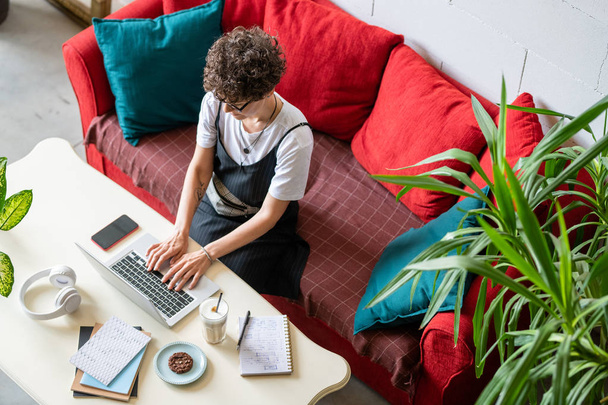 Casual młoda kobieta siedzi na kanapie przed laptopem, wpisując i surfując w sieci podczas pracy zdalnie - Zdjęcie, obraz