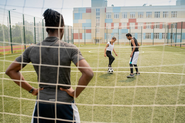 Takanäkymä nuori afrikkalainen urheilija seisoo net ja katsot kaksi pelaajaa jalkapallo kentällä
 - Valokuva, kuva
