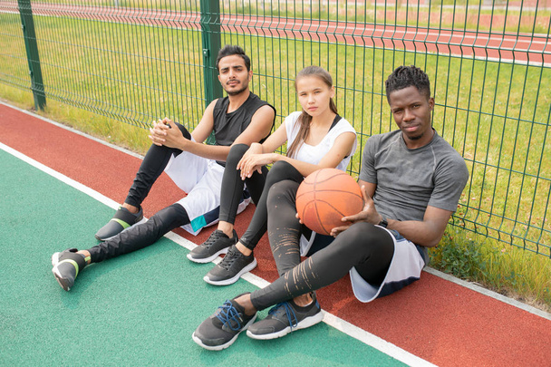 Tres jóvenes atletas en ropa deportiva sentados en cancha de baloncesto al aire libre por red y descansando después del partido
 - Foto, Imagen