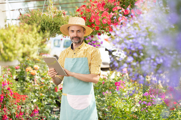 Dojrzały Brodaty ogrodnik w odzieży roboczej patrząc na Ciebie podczas poszukiwania informacji online na temat sadzenia - Zdjęcie, obraz
