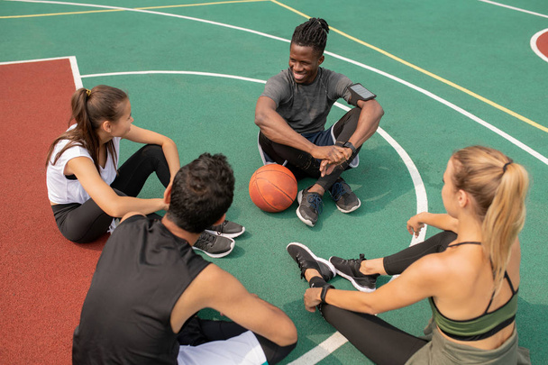 Group of young friendly intercultural people sitting on playground while getting ready for another game of basketball - Fotó, kép