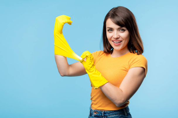Feliz joven ama de casa en camiseta amarilla ponerse guantes de goma en las manos antes de hacer las tareas domésticas
 - Foto, Imagen
