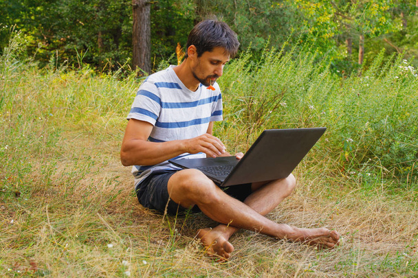 A young man sits on the grass with a flower in his mouth and works correspondence on a laptop. - Фото, изображение