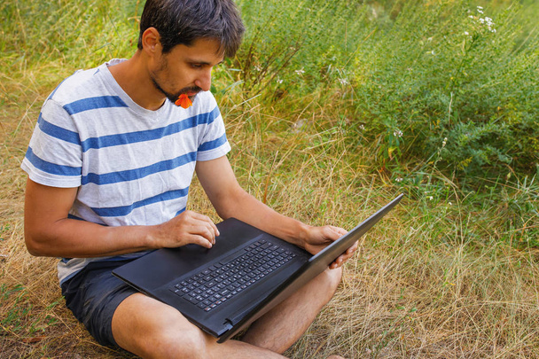 Een jonge man zit op het gras met een bloem in zijn mond en werkt correspondentie op een laptop. - Foto, afbeelding