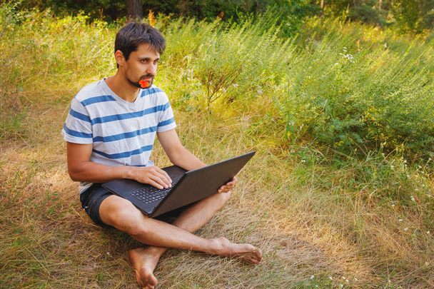 A young man sits on the grass with a flower in his mouth and works correspondence on a laptop. - Foto, Imagen