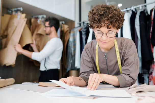 junge Näherin oder Modedesignerin betrachtet Skizze eines neuen Modells nach Arbeitsplatz im Atelier - Foto, Bild
