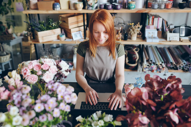 Content pretty redhead florist in apron standing at counter with flowers and working with social media of flower shop - Photo, Image