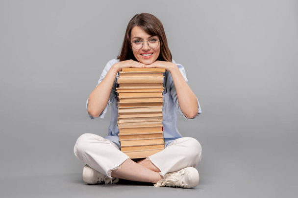 Νέα χαμογελαστή γυναίκα μαθητής με το πηγούνι της πάνω από μεγάλη στοίβα βιβλίων για να διαβάσει κοιτάζοντας σας σε απομόνωση - Φωτογραφία, εικόνα