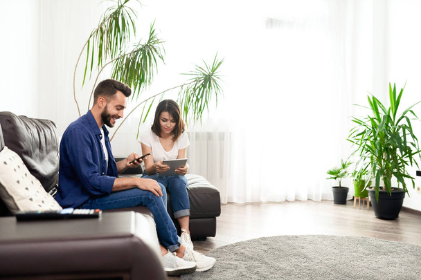 Позитивная молодая пара в повседневных нарядах сидит на удобном диване в квартире с Wi-Fi и с использованием устройств вместо связи
 - Фото, изображение
