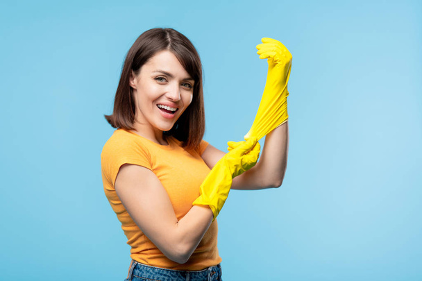 Η νεαρή οικονόμος στο καθημερινό ντύσιμο σας κοιτάζει με χαμόγελο ενώ βάζει κίτρινα λαστιχένια γάντια πριν από τις οικιακές εργασίες - Φωτογραφία, εικόνα