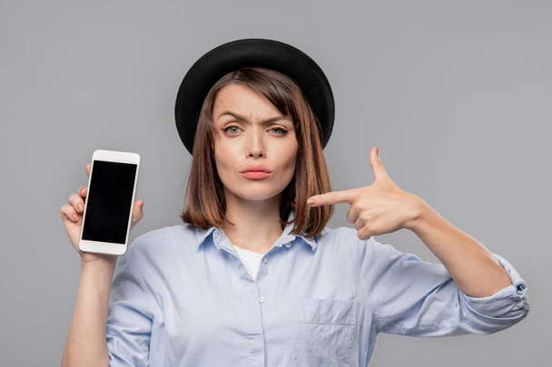 Brunette girl in hat and shirt showing you notification in smartphone while pointing at it in isolation - Photo, image