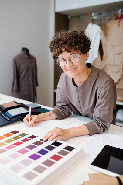 Joven diseñador alegre de ropa haciendo notas y mirando a través del catálogo de muestras textiles en el taller
 - Foto, Imagen