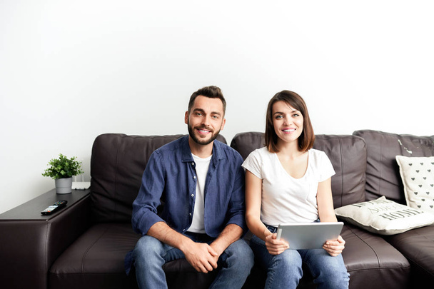 Portret szczęśliwej młodej pary w casual stroje siedzi na wygodnej kanapie w domu i za pomocą cyfrowego tabletu do oglądania filmu - Zdjęcie, obraz