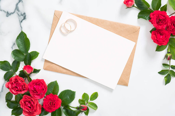 Mockup wedding invitation card or love letter with craft paper envelope, rose flowers, bridal rings on marble background. Minimal flat lay style composition, top view, overhead. - Foto, Imagen