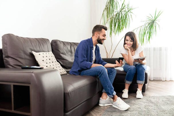lächelnder hübscher junger Mann zeigt Smartphone-Bildschirm, während er Internet-Nachrichten mit Freundin zu Hause teilt - Foto, Bild