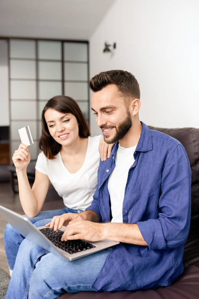 Весела молода пара сидить на дивані у вітальні і за допомогою кредитної картки і ноутбука при оплаті онлайн для замовлення - Фото, зображення