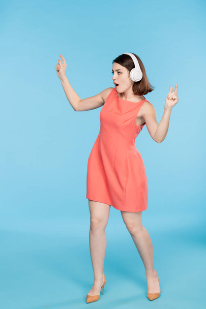Jovem mulher animada em elegante vestido vermelho e fones de ouvido desfrutando de música, cantando e dançando de forma isolada
 - Foto, Imagem