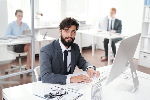 Retrato de hombre de negocios barbudo guapo sonriendo a la cámara mientras está sentado en el escritorio en la oficina, espacio de copia
 - Foto, Imagen