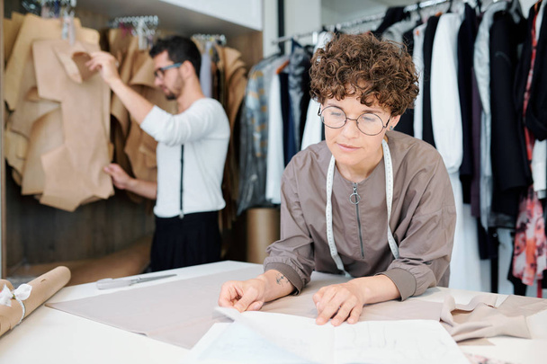 Νέοι σίγουροι σχεδιαστής μόδας κοιτάζοντας σκίτσο σε χαρτί, ενώ εργάζονται πάνω από τα νέα μοντέλα από το χώρο εργασίας - Φωτογραφία, εικόνα