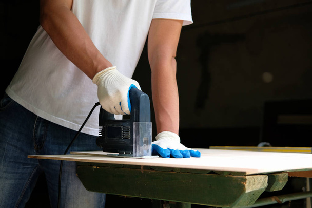 Carpenter in gloves using electric jig saw in workshop. Side view. Carpentry concept. - Φωτογραφία, εικόνα