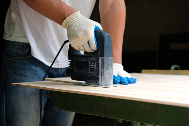 Carpenter in gloves using jig saw in workshop. Side view. - 写真・画像