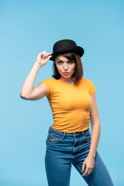 Mujer casual joven en sombrero negro, camiseta amarilla y vaqueros azules mirándote con expresión coqueta
 - Foto, Imagen