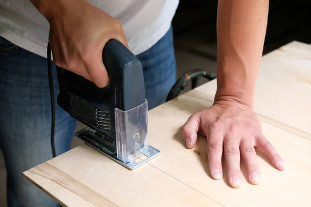 Tischler sägt in Tischlerei eine Sperrholzplatte mit elektrischer Stichsäge. hautnah Hand in Hand mit Puzzle. Zimmerei-Konzept. - Foto, Bild