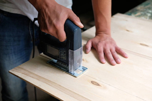 Carpenter is sawing a plywood sheet with electric jig saw machine in carpentry workshop. Close up hand with jigsaw. Carpentry concept. - Φωτογραφία, εικόνα