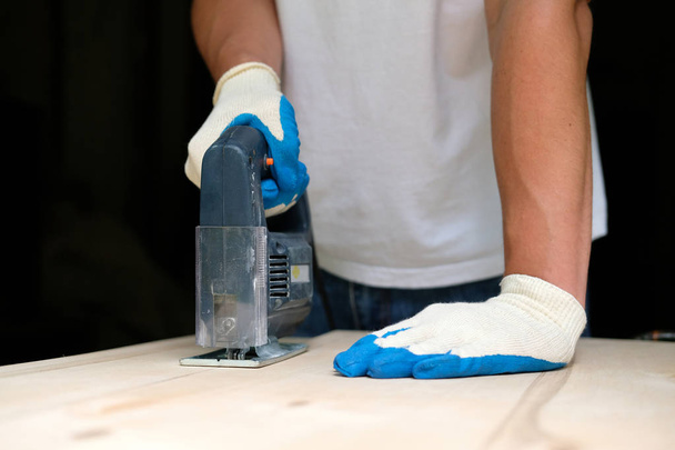 Carpenter in gloves using electric jig saw in workshop. Side view. Carpentry concept. - Foto, Bild