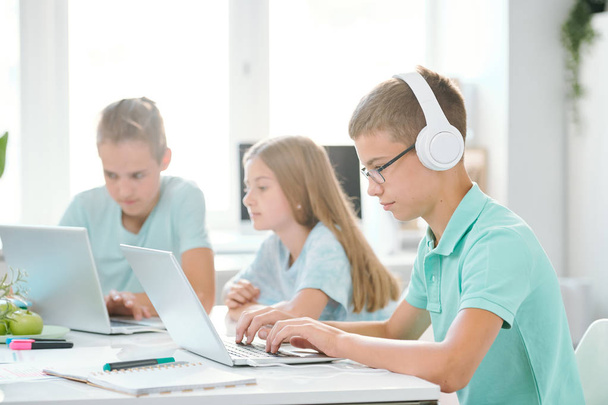 Un des jeunes camarades de classe dans les écouteurs regardant l'affichage de l'ordinateur portable tout en regardant la vidéo en ligne par bureau
 - Photo, image