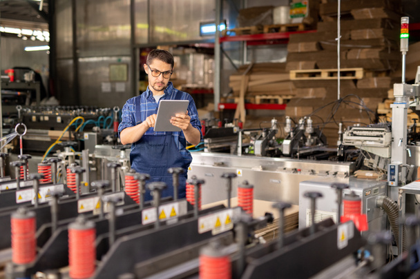 zeitgenössischer Ingenieur einer Industrieanlage mit Touchpad im Stehen an einer großen Industriemaschine in der Werkstatt - Foto, Bild