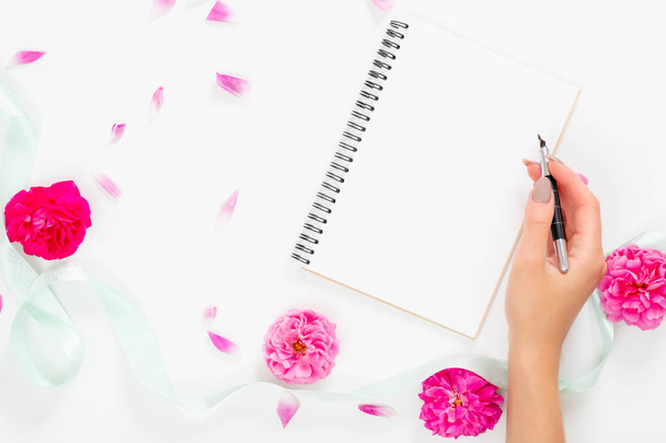 Female hand write a message in paper notebook lying on feminine desk with pink rose flowers and ribbon. Flatlay of woman home office desk table, top view. - Foto, Bild