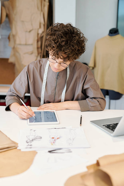 Досить молода серйозна жінка творчий дизайнер прокручування через цифрові ескізи в тачпад, сидячи за столом в майстерні
 - Фото, зображення
