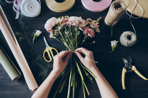 Vista de ángulo alto de florista irreconocible de pie junto a una mesa de madera con artículos de embalaje de ramo y atando flores con cuerda
 - Foto, Imagen
