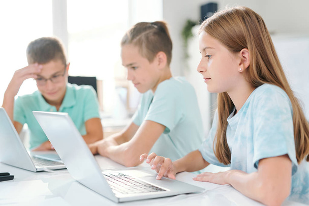Молодая серьезная школьница и ее одноклассники работают индивидуально перед ноутбуками, сидя за столом
 - Фото, изображение