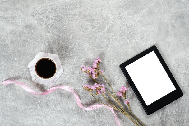 Flat lay elegant feminine accessories: coffee cup, pink ribbon, dry flowers and e book reader on concrete surface. Top view beauty blogger workspace, freelancer home office desk. Fashion blog hero. - 写真・画像