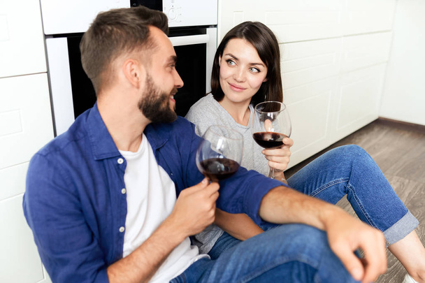 Ευτυχισμένο όμορφο νεαρό ζευγάρι σε casual ρούχα κάθονται στο πάτωμα και πίνοντας κόκκινο κρασί, ενώ γιορτάζει την επέτειο στο σπίτι - Φωτογραφία, εικόνα