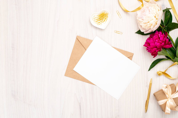 Flat lay home office desk. Female workspace with blank paper card, craft envelope, bouquet of peony flowers, golden accessories on wooden background. Top view feminine background. Fashion blog banner - Foto, imagen