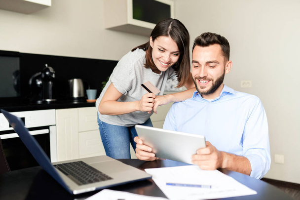 Positive excited young couple using digital tablet while choosing goods on internet store together in kitchen - Foto, Bild