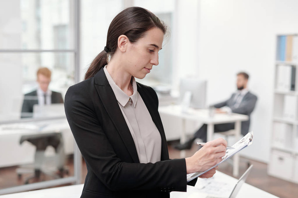 Retrato de vista lateral de una joven empresaria escribiendo en el portapapeles mientras está de pie en la oficina, espacio para copiar
 - Foto, Imagen