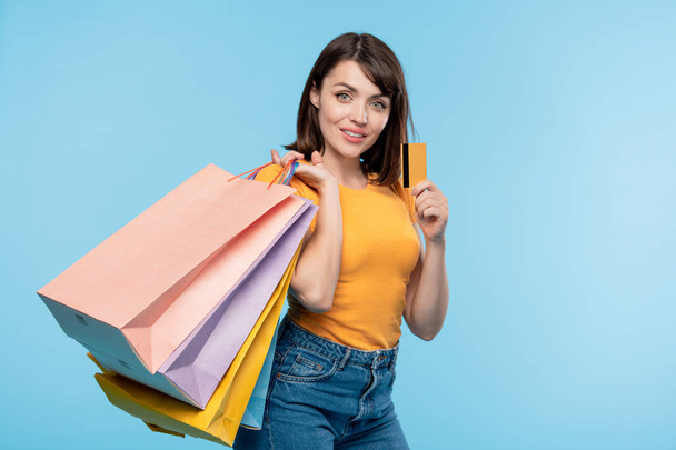 Glücklicher junger Shopaholic mit mehreren Paperbags prahlt nach erfolgreichem Einkauf mit gelber Kreditkarte - Foto, Bild