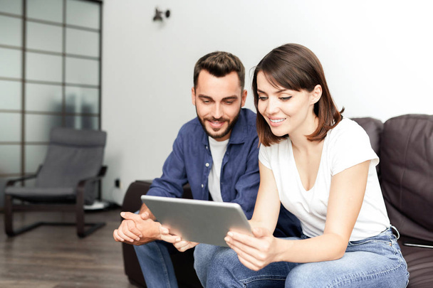 Sorrindo jovem casal sentado em sofá confortável na sala de estar e usando tablet para navegar na internet
 - Foto, Imagem