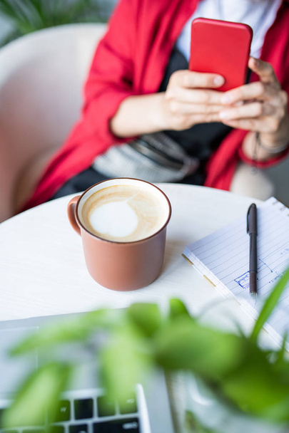 Mulher contemporânea com smartphone sentado à mesa no café e tirando foto de caneca de cappuccino
 - Foto, Imagem