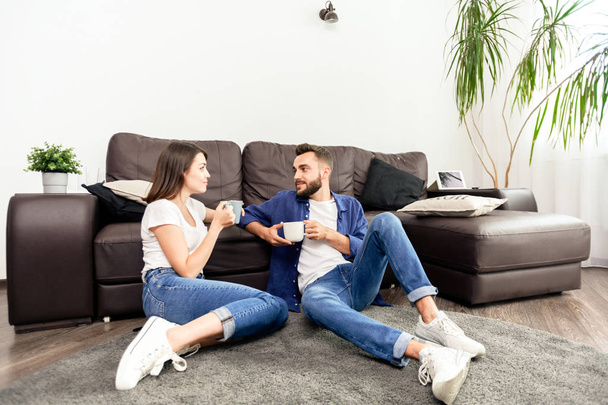 Contenuto giovane coppia in jeans e scarpe bianche seduti su moquette e bere caffè mentre trascorrete del tempo insieme a casa
 - Foto, immagini
