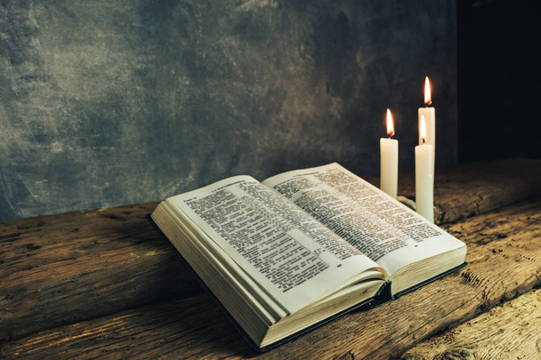 Kaunis avoin Raamattu ja kolme valkoista palanutta kynttilää vanhalla tammen puupöydällä ja tummanharmaa seinä tausta
.  - Valokuva, kuva