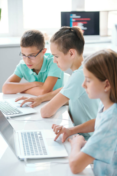 Rząd poważnych cute szkolnych dzieci w casualwear siedzi na biurku przed laptopami podczas przygotowań do seminarium szkolnego w przerwie - Zdjęcie, obraz