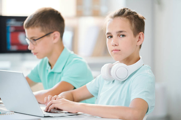 Cute and clever schoolboy with headpones on neck working over homework or presentation in front of laptop - Photo, Image